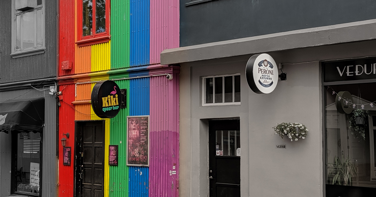 A photo of buildings in Iceland, it is edited to be black-and-white except for the queer bar which is painted in the colors of pride.